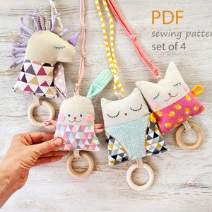 May include: Set of four handmade baby toys, including a unicorn, bunny, and two cat-shaped designs. Each toy features a wooden ring and is suspended by a colorful fabric cord. The toys are made with various patterned fabrics and soft textures. The text "PDF sewing pattern set of 4" is visible.