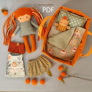 Peut inclure: Une poupée faite à la main avec des cheveux orange et une robe grise, ainsi qu'un lit, une couverture, un oreiller et des accessoires pour poupée. Le lit est orange avec un oreiller à pois et une couverture avec des illustrations de chats. Le texte "sewing pattern PDF" est inclus.