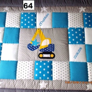 Peut inclure: Une courtepointe bébé en patchwork bleu et blanc avec un motif d'excavatrice jaune au centre. La courtepointe porte le nom "Mattias" et les dates "23.06.2016" et "19.09.2016" brodées dessus.