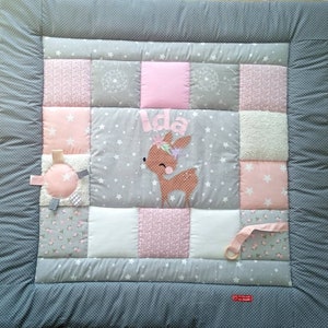 Può includere: Un tappeto gioco per bambini patchwork con un motivo a stelle grigie e bianche, accentuato da quadrati rosa e stampe floreali. Presenta un'applicazione di cervo con il nome "Ida" e giocattoli attaccati. Il bordo è grigio unito con pois bianchi.