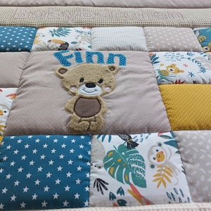 Puede incluir: Una manta de bebé beige con un diseño de patchwork. La manta presenta un oso de peluche marrón con el nombre "Finn" bordado en ella. La manta también tiene una variedad de otros patrones, incluyendo estrellas, hojas y animales.