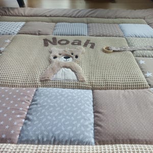 May include: A quilted play mat featuring a central beige waffle-textured panel with an embroidered brown bear and the name Noah. The mat is composed of various square patches in shades of beige, gray, and white, decorated with star and dot patterns.
