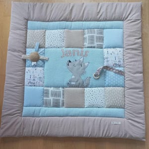 Op de afbeelding: Baby speelmat met een patchwork ontwerp in blauw, grijs en beige tinten. De mat heeft de naam "Janis" en een cartoon wolf. Inclusief bevestigde speeltjes en een gewatteerde rand.