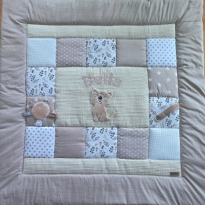 Peut inclure: Un tapis de jeu pour bébé en patchwork beige et blanc avec une applique de chat marron et le nom "Bella" brodé au centre.