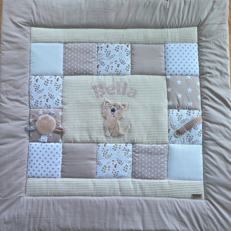 May include: A beige and white patchwork baby play mat with a brown cat applique and the name "Bella" embroidered in the center.