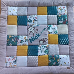 Könnte beinhalten: Ein Patchwork-Babyquilt mit verschiedenen quadratischen Mustern in Grün-, Gelb-, Grau- und Weißtönen. In der Mitte befindet sich ein graues Quadrat mit dem Namen "Nick" und Affenillustrationen. Das Datum "29.03.2024" ist ebenfalls sichtbar.