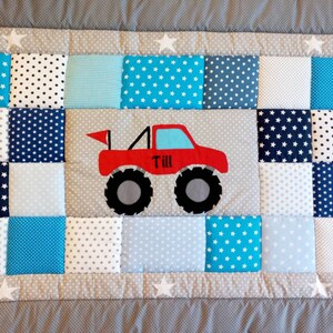 Peut inclure: Une courtepointe patchwork avec une application de camion monstre rouge. La courtepointe présente des carrés dans différentes nuances de bleu, blanc et gris avec des motifs d'étoiles et de pois. Le nom "Till" est sur le camion.