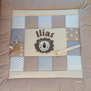 Puede incluir: Una alfombra de juego para bebés de patchwork con un diseño de león y el nombre "Ilias". La alfombra presenta varias telas en tonos neutros, incluyendo estrellas, lunares y patrones de cuadros. Un pequeño juguete y una cinta están adjuntos.