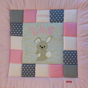 Op de afbeelding: Een roze en grijze babydeken met een konijn en de naam "Lea" erop geborduurd. De deken heeft een patchwork-ontwerp met verschillende stoffen, waaronder ruitjes, sterren en stippen. De datum "13.08.2020" is ook op de deken geborduurd.