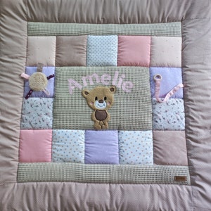 Op de afbeelding: Een beige babydeken met een patchwork ontwerp met roze, witte en paarse vierkanten. De deken heeft een bruine teddybeer-applicatie en de naam "Amelie" geborduurd in roze.