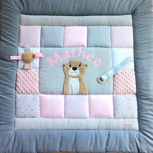 Op de afbeelding: Een vierkante patchwork speelmat met een donkergrijze rand met stippen, lichtgrijze wafelstof en roze textuurvierkanten. Het midden toont een geborduurde beige otter en de naam Mathea in roze letters, met aangehechte stoffen lussen en een houten ring.