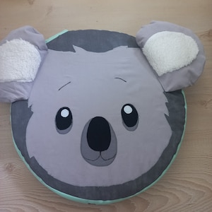 Könnte beinhalten: Ein graues und weißes Plüschkissen in Koalaform mit einem hellgrünen Saum. Das Kissen hat gestickte Augen, eine Nase und einen Mund.