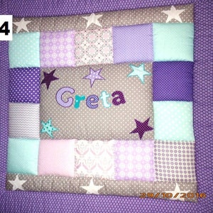 Könnte beinhalten: Eine lila und graue Patchwork-Decke mit einem hellblauen, rosa und weißen Muster. Die Decke hat einen Namen, "Greta", der in der Mitte gestickt ist. Die Decke ist mit Sternen verziert.