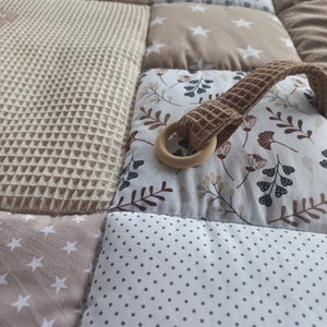 May include: A beige and brown baby play mat with a variety of textures and patterns. The mat features a wooden ring attached to a brown fabric loop.