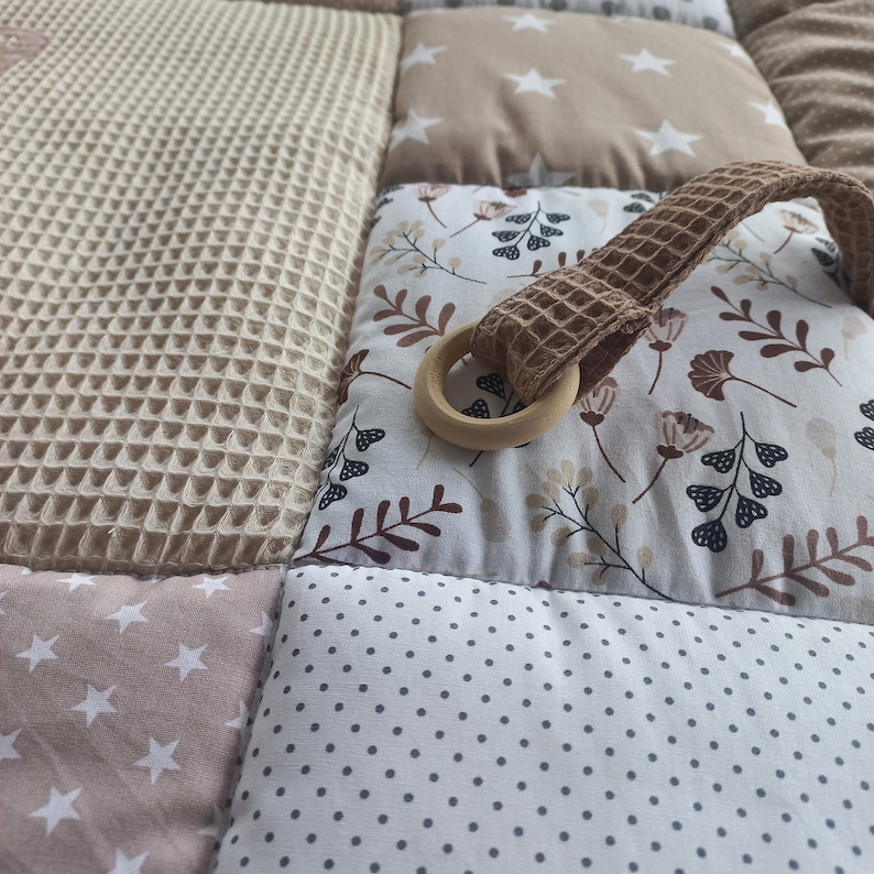 May include: A beige and brown baby play mat with a variety of textures and patterns. The mat features a wooden ring attached to a brown fabric loop.
