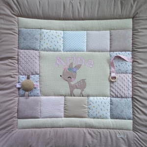 Könnte beinhalten: Eine Baby-Spielmatte mit Patchwork-Design in Rosa-, Beige- und Weißtönen. Die Matte zeigt eine Hirschapplikation mit dem Namen "Arina" darüber gestickt. Enthält einen Holzring und ein strukturiertes Blumenspielzeug.