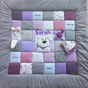 Op de afbeelding: Een paarse, grijze en witte babydeken met een witte pluche hond-applique. De deken heeft een verscheidenheid aan patronen, waaronder sterren, stippen en bloemenpatronen. De deken bevat ook een label met de tekst "3460 g" en een label met de tekst "27.02.24".