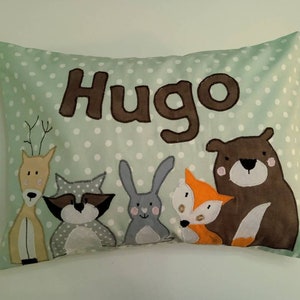 Op de afbeelding: Een groen kussen met witte stippen en de naam "Hugo" geborduurd in bruin. Het kussen heeft vijf aangebrachte dierengezichten: een hert, een wasbeer, een konijn, een vos en een beer.