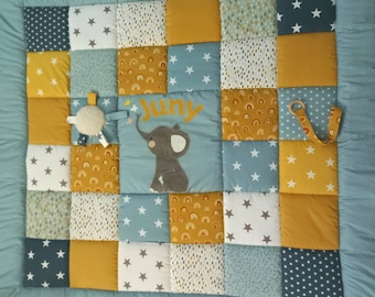 Peut inclure: Un tapis d'éveil bébé patchwork bleu et jaune avec un éléphant cousu et le nom "Juni" dessus. Le tapis d'éveil a une variété de textures et de motifs, y compris des étoiles, des pois et des arcs-en-ciel.