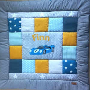 Könnte beinhalten: Eine Patchwork-Babydecke mit grauem Polka-Dot-Rand und weißen Sternakzenten. Die Mitte zeigt den Namen "Finn" in Gelb, ein blaues Rennauto und Quadrate aus blauen, gelben und weißen Stoffen. Die Decke misst ca. 76 x 76 cm.