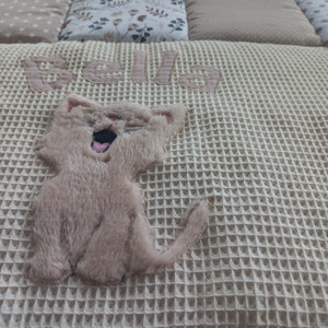May include: A cream-colored waffle weave blanket with the name "Bella" embroidered in pink and a brown plush cat applique.