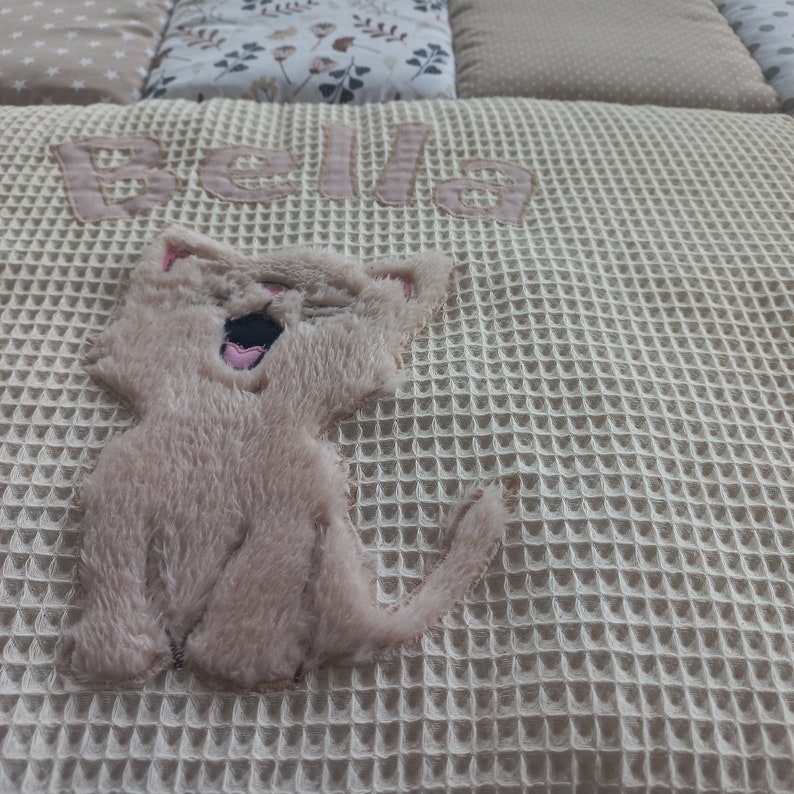 May include: A cream-colored waffle weave blanket with the name "Bella" embroidered in pink and a brown plush cat applique.