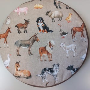 Op de afbeelding: Rond stoffen item met illustraties van boerderijdieren. Het ontwerp bevat illustraties van een varken, schaap, hond, eend, paard, ezel en andere dieren. De achtergrond is een neutrale beige kleur en de rand is donkerbruin.
