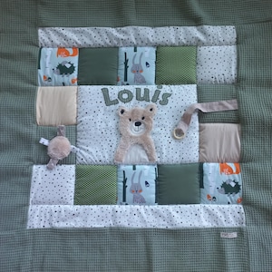 Può includere: Una coperta patchwork quadrata per neonati con un bordo verde oliva a trama waffle. Il centro presenta un'applicazione di orsetto di peluche beige, il nome Louis ricamato e vari quadrati di tessuto con stampe di animali, pois e colori a tinta unita.