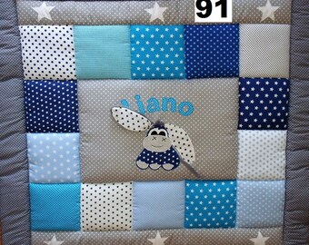 Peut inclure: Une couverture patchwork pour bébé aux tons de bleu, blanc et gris, avec un motif central d'un âne de dessin animé et le nom "Iano". La couverture est bordée de tissu gris et d'étoiles blanches.