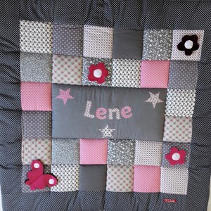 Peut inclure: Une couverture bébé en patchwork gris et rose avec le nom "Lene" brodé au centre. La couverture présente différents motifs, notamment des étoiles, des fleurs et des papillons.