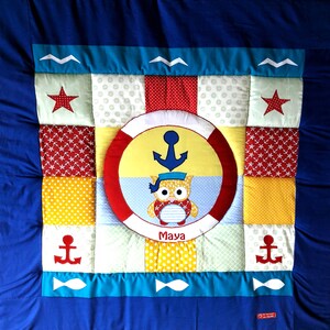 May include: A colourful patchwork quilt with a nautical theme. The centre features a yellow circle with a red life preserver around it. Inside the circle is a blue anchor and a cartoon owl wearing a sailor hat. The quilt is made up of various fabrics with different patterns, including stars, anchors, and fish. The quilt is personalised with the name "Maya".