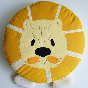 Op de afbeelding: Een rond, geel en wit pluche kussen in de vorm van een leeuw met een witte vachtmanen en poten. Het gezicht van de leeuw is geborduurd met zwarte ogen, een zwarte neus en een glimlachende mond.
