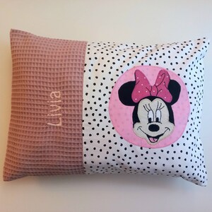 Könnte beinhalten: Ein rechteckiges Kissen mit einem rosa Waffelstoff auf der linken Seite und einem weißen Stoff mit schwarzen Punkten auf der rechten Seite. Es zeigt ein gesticktes Minnie Mouse-Design und den Namen "Livia".