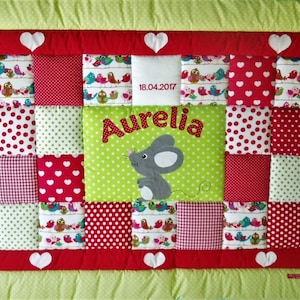 Op de afbeelding: Een groene, rode en witte patchwork babydeken met een muis-applicatie en de naam "Aurelia" geborduurd erop. De datum "18.04.2017" is ook geborduurd op de deken.