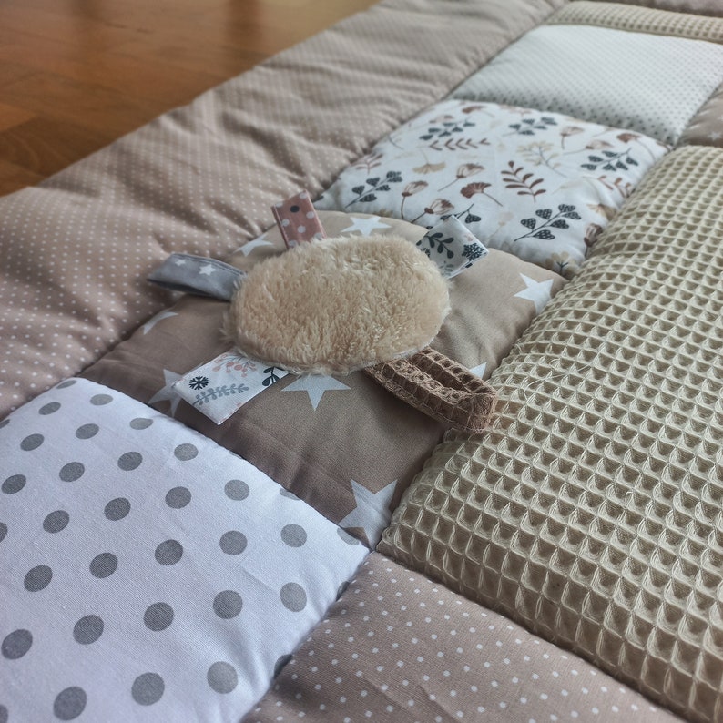 May include: A beige and brown patchwork play mat with a plush, beige toy attached. The mat is made of different textures, including polka dots, stars, and waffle weave.