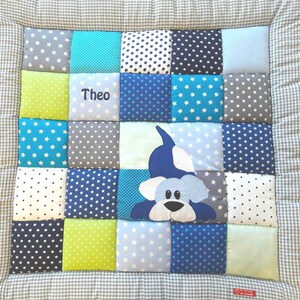 Op de afbeelding: Een blauwe en groene patchwork baby speelmat met een witte geborduurde naam "Theo" en een blauwe en witte applicatie van een hondenkop.