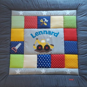 Peut inclure: Une courtepointe patchwork pour bébé avec une bordure grise et un panneau central avec le nom "Lennard" et une excavatrice jaune. La courtepointe comprend des carrés de différentes couleurs et motifs, dont des étoiles, des pois et du vichy.