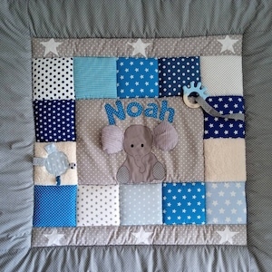 May include: A gray and blue baby play mat with a patchwork design. The mat features a gray elephant with a blue and white polka dot pattern, a blue and white star pattern, and a blue and white checkered pattern. The mat also includes a blue and white star-shaped rattle and the name "Noah" embroidered in the center.