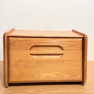 May include: A rectangular wooden bread box with a light brown natural wood grain finish. The front panel features a horizontal recessed handle cutout, and the unit has rounded side edges, providing a clean, functional design for kitchen storage.