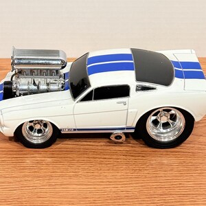 May include: A white toy car with blue racing stripes and chrome wheels. The car has a large engine and a black roof. The model is a GT 350.