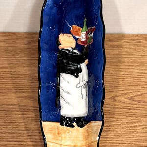 May include: A long, oval ceramic serving platter with a whimsical design. The platter features a blue background with a painted image of a waiter holding a tray with food and wine. The bottom portion is tan, and the edges are black.