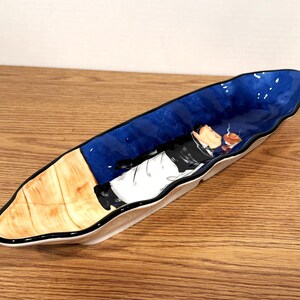 May include: A long, ceramic serving dish with a blue interior and a black rim. The dish features a painted illustration of a person in a suit, smoking a pipe. The exterior has a light brown and white design.