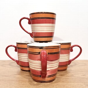 May include: Four ceramic mugs with a striped design in shades of orange, red, and white. Each mug has a curved handle and black accent lines. The mugs are stacked on a wooden surface.
