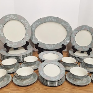 May include: A complete vintage dinnerware set with a light blue floral design. The set includes plates, saucers, bowls, and teacups. The plates are stacked on top of each other. The set is displayed on a wooden surface.