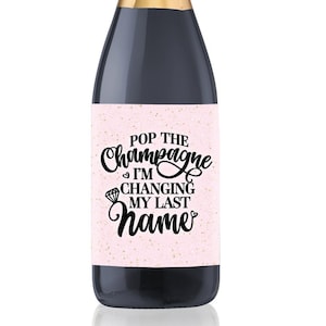 Könnte beinhalten: Eine Champagnerflasche mit einem rosa Etikett, auf dem "Pop the Champagne I'm Changing My Last Name" mit einer Diamant-Ring-Grafik und goldenem Glitzer steht.