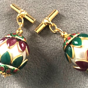 May include: A pair of gold-toned cufflinks with an ornate design. Each cufflink features a white, egg-shaped center surrounded by green and burgundy accents. The design is further embellished with red and green details.