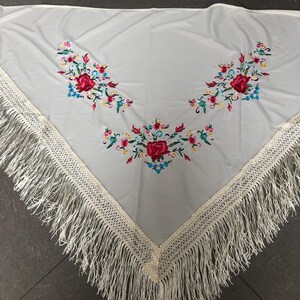 May include: White triangular shawl with embroidered floral designs in red, pink, blue, and yellow. The shawl has a cream-colored crocheted border and long fringe along the edges. The floral embroidery is arranged in three clusters.