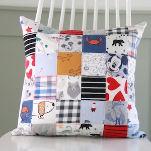 May include: A patchwork pillow with a variety of colorful fabrics featuring cartoon animals, characters, and patterns. The pillow is made from repurposed baby clothes.