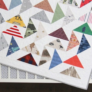 May include: A white baby quilt with a geometric pattern of colorful triangles. The quilt is made from various fabrics with different patterns, including stripes, stars, and animals. The quilt is lying on a white crib with black polka dots.
