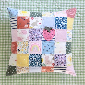 May include: A patchwork pillow with a variety of colorful fabrics featuring floral patterns, animals, and geometric shapes. The pillow is quilted with a curved design and has a pink strawberry applique in the center.